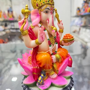 May include: A colourful statue of Ganesha, the Hindu god of wisdom and prosperity, seated on a pink lotus flower. He is holding a sweet treat in his left hand and a small axe in his right hand. A small grey mouse is sitting at the base of the lotus.