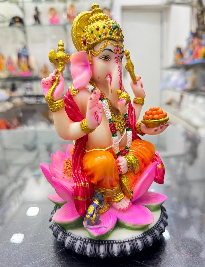 May include: A colourful statue of Ganesha, the Hindu god of wisdom and prosperity, seated on a pink lotus flower. He is holding a sweet treat in his left hand and a small axe in his right hand. A small grey mouse is sitting at the base of the lotus.