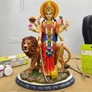 May include: A detailed statue of a multi-armed deity, adorned in red and blue garments, a golden crown, and jewelry. The figure stands with a lion, holding a sword and a golden mace. The base is intricately designed with a floral pattern.