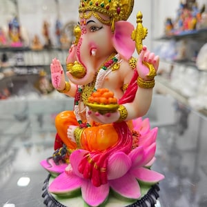 May include: A colourful figurine of the Hindu deity Ganesha, seated on a pink lotus flower. He is wearing a gold crown and a red and orange sari. He is holding a bowl of sweets in his right hand and a small mouse is at his feet.