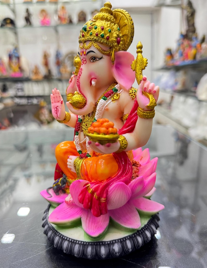 May include: A colourful figurine of the Hindu deity Ganesha, seated on a pink lotus flower. He is wearing a gold crown and a red and orange sari. He is holding a bowl of sweets in his right hand and a small mouse is at his feet.