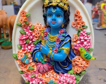 May include: A blue and gold figurine of Krishna, a Hindu deity, surrounded by colorful flowers. The figurine is set against a white oval background.