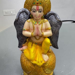 May include: A golden statue of a deity with wings, sitting on a golden base. The deity is wearing a yellow robe and has a golden crown. The statue is painted with intricate details and has a traditional Indian style.