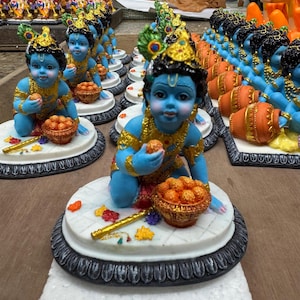 May include: A collection of small blue figurines of the Hindu god Krishna, each holding a bowl of yellow balls. The figurines are sitting on oval-shaped bases with intricate designs.