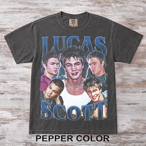May include: Pepper color t-shirt featuring a collage of images and the text "LUCAS SCOTT" in blue. The shirt has a vintage aesthetic, with a crew neck and short sleeves. The Comfort Colors brand tag is visible at the top.
