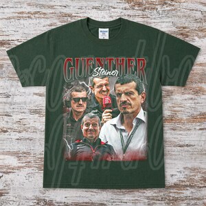 May include: Dark green t-shirt featuring a graphic of G&uuml;nther Steiner, with his name in red and white. The design includes multiple images of Steiner, with a lightning bolt effect in the background. The shirt is a Gildan brand.