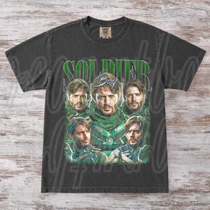 May include: Dark gray t-shirt featuring a graphic of a man in a green and black costume. The word "SOLDIER" is printed in green above the man's image, with the word "Boy" in a script font below.