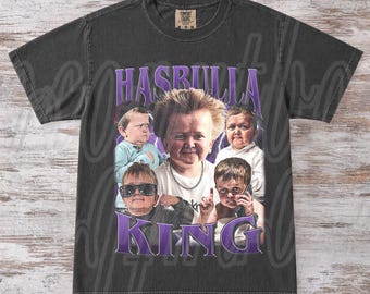 May include: A charcoal gray short-sleeve t-shirt featuring a graphic print of Hasbulla in various poses. The text "HASBULLA" appears in purple at the top and "KING" at the bottom. The shirt has a crew neckline and a vintage-style washed finish.