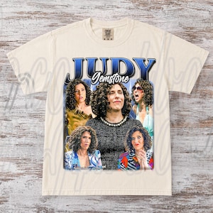 以下が含まれることがあります： クリーム色のTシャツ。青色の「JUDY」の文字と、その下に「Gemstone」のグラフィックがプリントされています。グラフィックには、巻き毛の女性の複数の画像が含まれています。シャツはComfort Colorsブランドです。