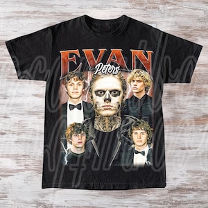 Może przedstawiać: Czarny t-shirt z kolażem zdjęć Evana Petersa. Projekt zawiera napis "EVAN Peters" w pomarańczowej i białej czcionce w stylu vintage. Centralny obraz przedstawia Petersa z makijażem czaszki.