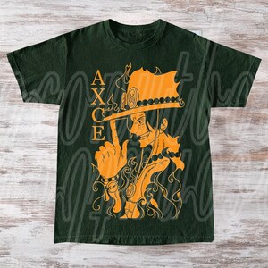 以下が含まれることがあります： オレンジ色のイラストが特徴のダークグリーンのTシャツ。帽子をかぶったキャラクターと「ACE」の文字が描かれています。キャラクターは指を指しています。デザインには炎とネックレスが含まれています。