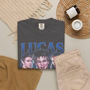 May include: Dark gray t-shirt with the name "LUCAS" in blue lettering and three faces. The shirt is paired with tan pants and a woven placemat. A candle and a woven coaster are also in the image.