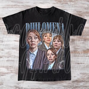 May include: Black t-shirt featuring a graphic of four images of a woman in a blazer and collared shirt. The text "PHILOMENA Cunk" is printed above the images in a blue, stylized font. Lightning bolt graphics are on the sides.