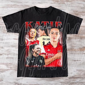 Könnte beinhalten: Schwarzes T-Shirt mit einer Collage von Bildern von Katie McCabe, mit dem Namen "Katie McCabe" in roter Schrift. Das Shirt enthält Bilder von McCabe in einem rot-weißem Fußballtrikot und einem gelben Trikot.