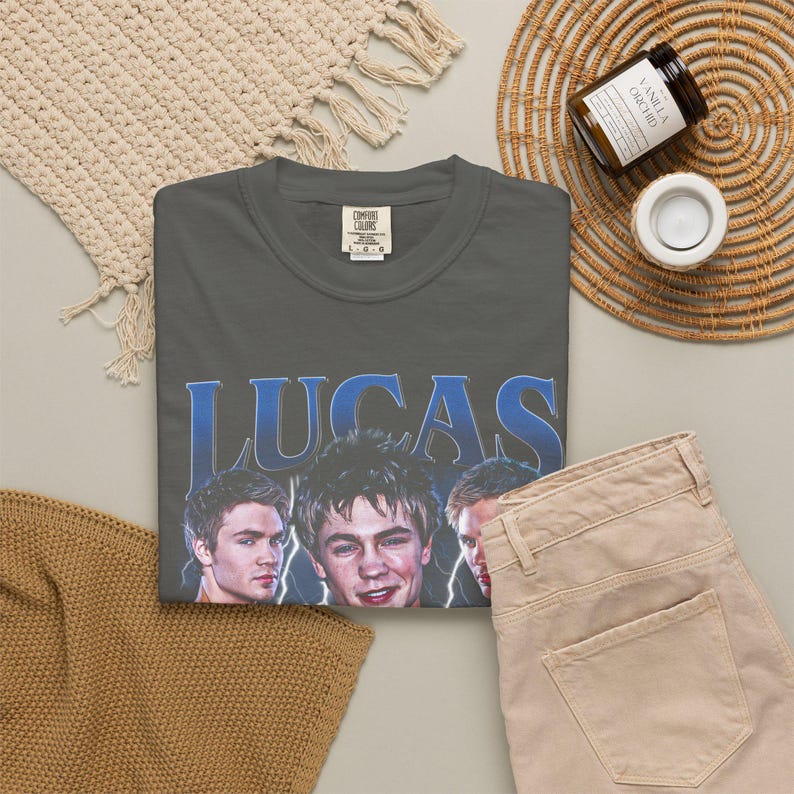 May include: A charcoal gray t-shirt with the name "LUCAS" in blue lettering and images of three men. The shirt is paired with tan pants, a woven placemat, a candle, and a woven coaster.