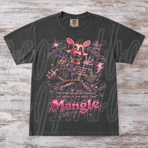 May include: Dark grey t-shirt with a graphic of a character with pink accents, stars, and chains. The text "Mangle" is in pink, with the quote "don't tell anyone but there all just jealous of how pretty I am."