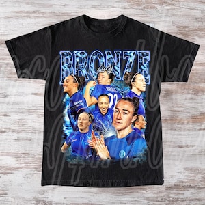 May include: Black t-shirt featuring a collage of images of a woman in a blue soccer uniform. The word "BRONZE" is displayed in a blue, stylized font above the images. The name "Lucy" is also visible.