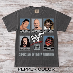 Pode incluir: Uma camiseta cinza escuro com retratos de seis lutadores da WWE: The Rock, Stone Cold, Mankind, The Undertaker e Kane. Cada retrato inclui detalhes de altura e peso. A camiseta também apresenta o texto "Superstars of the New Millennium".
