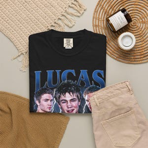 May include: Black t-shirt with the name "LUCAS" in blue, featuring images of three people. The shirt is paired with tan pants. A candle and woven mat are also visible.