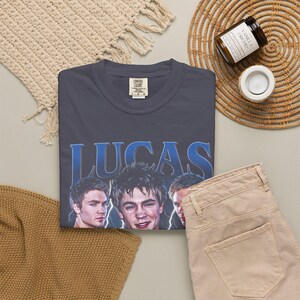 May include: A navy blue t-shirt with the name "LUCAS" in blue lettering and images of three men. The shirt is paired with tan pants and a woven mat. A candle and a woven basket are also in the image.