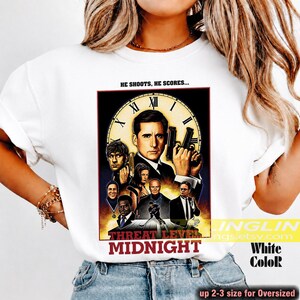 May include: A white t-shirt featuring a graphic print of a movie-style poster titled "Threat Level Midnight." The design includes a large clock face, multiple character portraits, and the text "HE SHOOTS, HE SCORES..." at the top of the rectangular image.