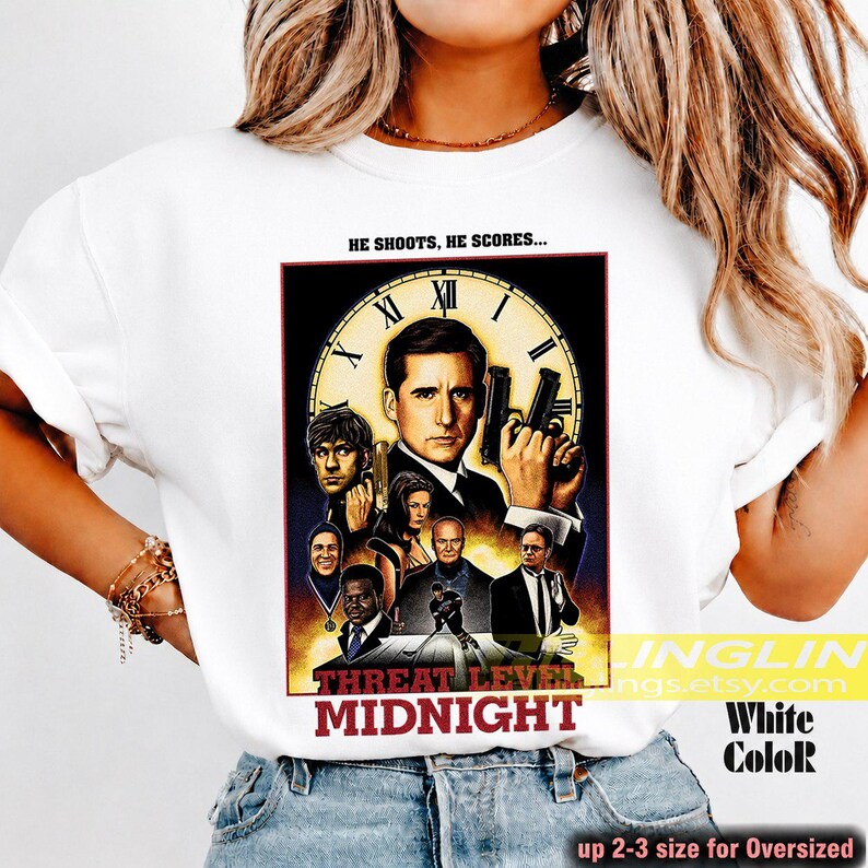 May include: A white t-shirt featuring a graphic print of a movie-style poster titled "Threat Level Midnight." The design includes a large clock face, multiple character portraits, and the text "HE SHOOTS, HE SCORES..." at the top of the rectangular image.
