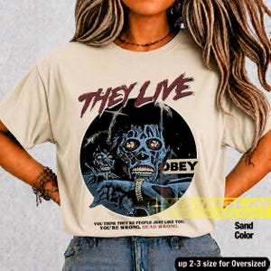 K&ouml;nnte beinhalten: Ein sandfarbenes T-Shirt mit einem Grafikdruck einer skelettartigen Figur und dem Text THEY LIVE sowie OBEY. Unter der Grafik steht der Text: YOU THINK THEY'RE PEOPLE JUST LIKE YOU. YOU'RE WRONG. DEAD WRONG. Das T-Shirt hat einen Rundhalsausschnitt.