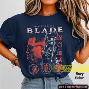 May include: A navy blue t-shirt featuring a graphic print of the film Blade. The design includes the text WESLEY SNIPES BLADE, a large portrait of the character, a figure holding a sword, and three circular character icons at the bottom.