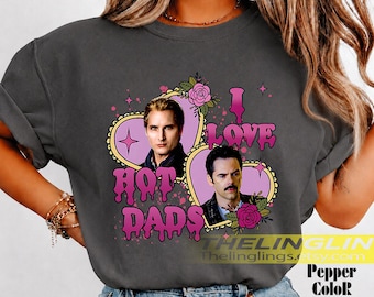 May include: A charcoal grey t-shirt featuring a graphic print of two men inside heart-shaped frames. The text reads I LOVE HOT DADS in a pink, dripping font. The design includes pink sparkles and pink roses. The shirt has short sleeves and a crew neck.