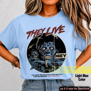 K&ouml;nnte beinhalten: Ein hellblaues T-Shirt mit einem Grafikdruck aus dem Film They Live. Das Design zeigt eine skelettartige Figur in einem schwarzen Kreis mit dem Text "THEY LIVE" dar&uuml;ber, "OBEY" auf einem Schild und dem Zitat "YOU THINK THEY'RE PEOPLE JUST LIKE YOU. YOU'RE WRONG. DEAD WRONG."
