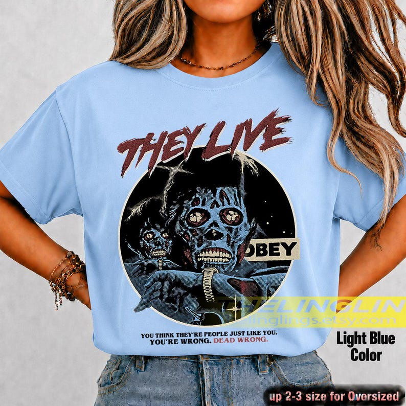 K&ouml;nnte beinhalten: Ein hellblaues T-Shirt mit einem Grafikdruck aus dem Film They Live. Das Design zeigt eine skelettartige Figur in einem schwarzen Kreis mit dem Text "THEY LIVE" dar&uuml;ber, "OBEY" auf einem Schild und dem Zitat "YOU THINK THEY'RE PEOPLE JUST LIKE YOU. YOU'RE WRONG. DEAD WRONG."