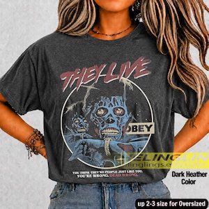 K&ouml;nnte beinhalten: Ein dunkelgrau meliertes T-Shirt mit einem Grafikdruck einer skelettartigen Figur und dem Text "THEY LIVE" in Rot &uuml;ber einem kreisf&ouml;rmigen Rahmen. Das Design enth&auml;lt das Wort "OBEY" sowie den Text "YOU THINK YOU ARE PEOPLE JUST LIKE YOU. YOU ARE WRONG. DEAD WRONG."