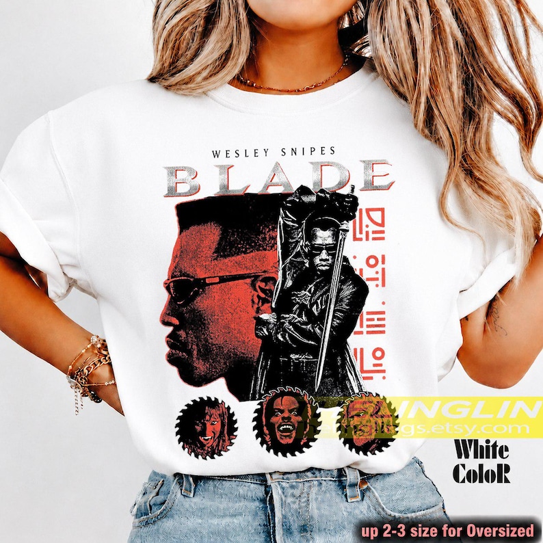 May include: A white t-shirt featuring a graphic print of Wesley Snipes as Blade. The design includes a large profile portrait, a full-body figure holding a sword, and three circular character icons at the bottom with red text accents.