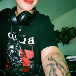 May include: A black t-shirt featuring a graphic print of the character Blade with red accents and the text "WESLEY SNIPES" and "BLADE" in white. The shirt is worn with black over-ear headphones and a gold necklace, with a visible arm tattoo.