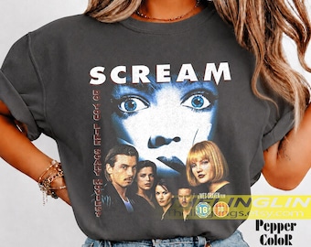 May include: A charcoal gray short-sleeve t-shirt featuring a graphic print from the movie Scream. The design includes the film title, a large blue-toned eye illustration, and a cast collage with the text Do You Like Scary Movies?
