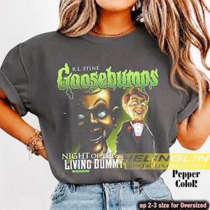 Op de afbeelding: Een antracietgrijs t-shirt met een grafische print van de Goosebumps-boekomslag Night of the Living Dummy. Het ontwerp bevat een grote, gedetailleerde illustratie van een buikspreekpop met groene ogen en de tekst R.L. Stine Goosebumps.