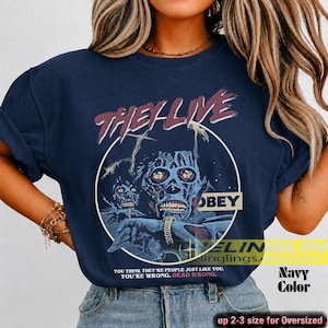 K&ouml;nnte beinhalten: Ein marineblaues T-Shirt mit einem Grafikdruck einer skelettierten Figur und dem Text THEY LIVE sowie OBEY. Unter der Grafik steht der Text: YOU THINK THEY ARE PEOPLE JUST LIKE YOU. YOU ARE WRONG. DEAD WRONG. Das Shirt hat kurze &Auml;rmel und einen Rundhalsausschnitt.