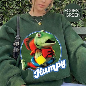 May include: Forest green sweatshirt with a cartoon fish graphic. The graphic features a green fish with red and orange accents, wearing a life preserver and a yellow square. The word "Humpy" is written in white script below the fish.