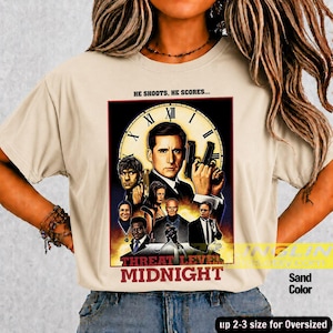 May include: A sand-colored t-shirt featuring a graphic print of a movie poster titled "Threat Level Midnight." The design includes a large clock face, multiple character portraits, and the text "HE SHOOTS, HE SCORES..." above the main title.