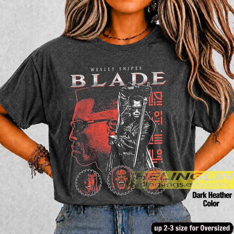 May include: A dark heather grey t-shirt featuring a graphic print of the film Blade. The design includes the text Wesley Snipes Blade, a large illustration of the character holding a sword, and three circular portraits at the bottom.