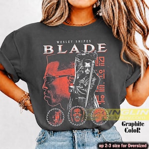 May include: A charcoal grey t-shirt featuring a red and white graphic print of Wesley Snipes as Blade holding a sword. The design includes the text WESLEY SNIPES BLADE, three circular saw blade icons with faces, and geometric patterns.
