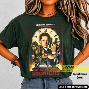 May include: A forest green t-shirt featuring a graphic print of the movie poster for Threat Level Midnight. The design includes a large clock face, multiple characters from the film, and the text HE SHOOTS, HE SCORES... and THREAT LEVEL MIDNIGHT.