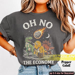 Pode incluir: Uma t-shirt cinzento-carvão com um gráfico de três dinossauros a usar gravatas. O texto diz OH NO THE ECONOMY. Um meteoro em chamas é representado acima dos dinossauros, um dos quais transporta uma pasta. O design tem um aspeto desgastado.
