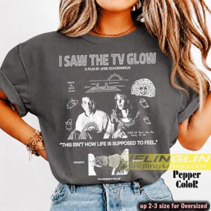 Op de afbeelding: Een antracietgrijs t-shirt met witte grafische prints, waaronder de tekst I SAW THE TV GLOW, A FILM BY JANE SCHOENBRUN en THIS IS HOW LIFE IS SUPPOSED TO FEEL. Het ontwerp bevat illustraties van twee personen, een zon en geesten.