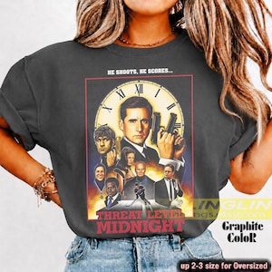 May include: A charcoal gray t-shirt featuring a graphic print of the movie poster for Threat Level Midnight. The design includes a large clock face, a central male figure holding a gun, and several smaller character portraits. The text reads HE SHOOTS, HE SCORES... and THREAT LEVEL MIDNIGHT.
