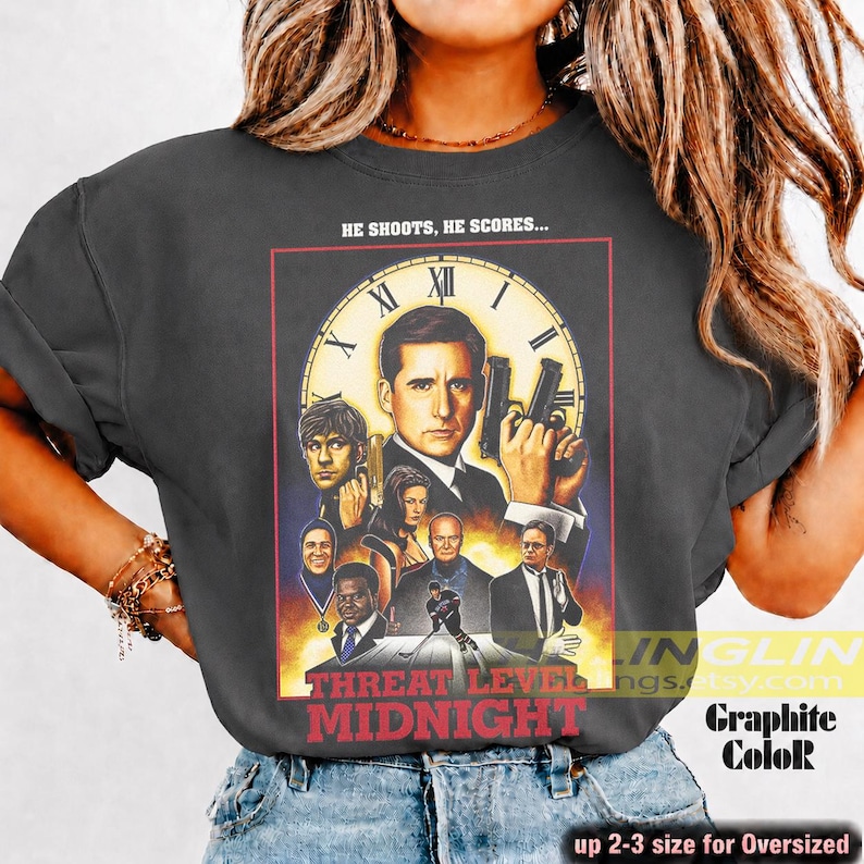 May include: A charcoal gray t-shirt featuring a graphic print of the movie poster for Threat Level Midnight. The design includes a large clock face, a central male figure holding a gun, and several smaller character portraits. The text reads HE SHOOTS, HE SCORES... and THREAT LEVEL MIDNIGHT.