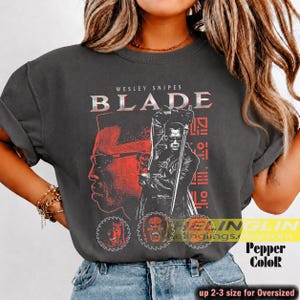 Może przedstawiać: Ciemnoszary t-shirt z nadrukiem graficznym przedstawiającym Wesleya Snipesa jako Blade'a. Projekt zawiera biały napis "WESLEY SNIPES BLADE", czerwoną sylwetkę twarzy oraz centralną postać trzymającą miecz z czerwonymi geometrycznymi akcentami.