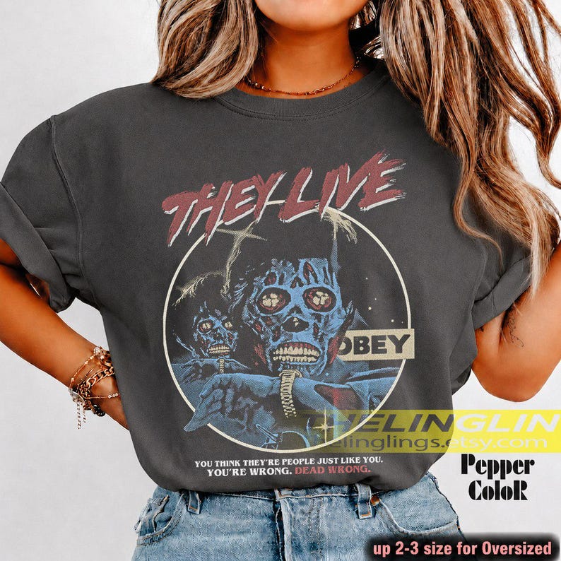 K&ouml;nnte beinhalten: Ein anthrazitfarbenes T-Shirt mit einem Grafikdruck aus dem Film They Live. Das Design zeigt eine blaue Alien-Figur, den Text "THEY LIVE" in Rot, "OBEY" in einem gelben Kasten und das Zitat "YOU THINK THEY'RE PEOPLE JUST LIKE YOU. YOU'RE WRONG. DEAD WRONG."
