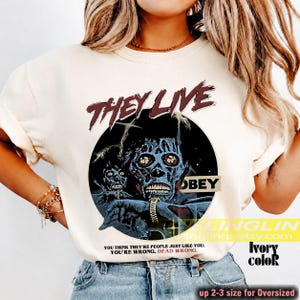 K&ouml;nnte beinhalten: Ein elfenbeinfarbenes T-Shirt mit einem Grafikdruck aus dem Film They Live. Das Design enth&auml;lt den Text THEY LIVE in Dunkelrot, eine kreisf&ouml;rmige Illustration einer Alien-Figur, das Wort OBEY und den Text YOU THINK THEY'RE PEOPLE JUST LIKE YOU. YOU'RE WRONG. DEAD WRONG.