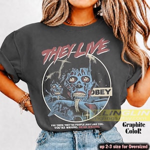 K&ouml;nnte beinhalten: Ein anthrazitfarbenes Kurzarm-T-Shirt mit einem Grafikdruck von zwei skelettierten Figuren in einem Kreis. Der Text lautet "THEY LIVE" oben, "OBEY" rechts und "YOU THINK THEY'RE PEOPLE JUST LIKE YOU. YOU'RE WRONG. DEAD WRONG." am unteren Rand.
